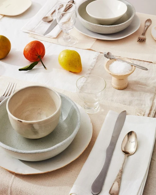 Overhead shot of a table setting with bowls, plates, utensils, glasses, and assorted citrus fruits arranged on a textured tablecloth.