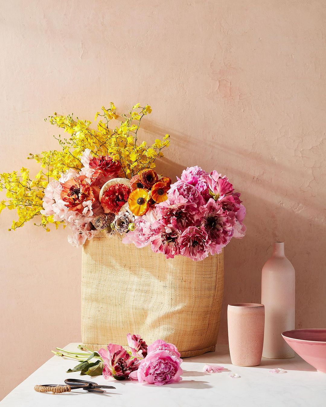 Floral arrangement with pink and yellow flowers in a straw bag, presented on a white table against a textured pink wall background.