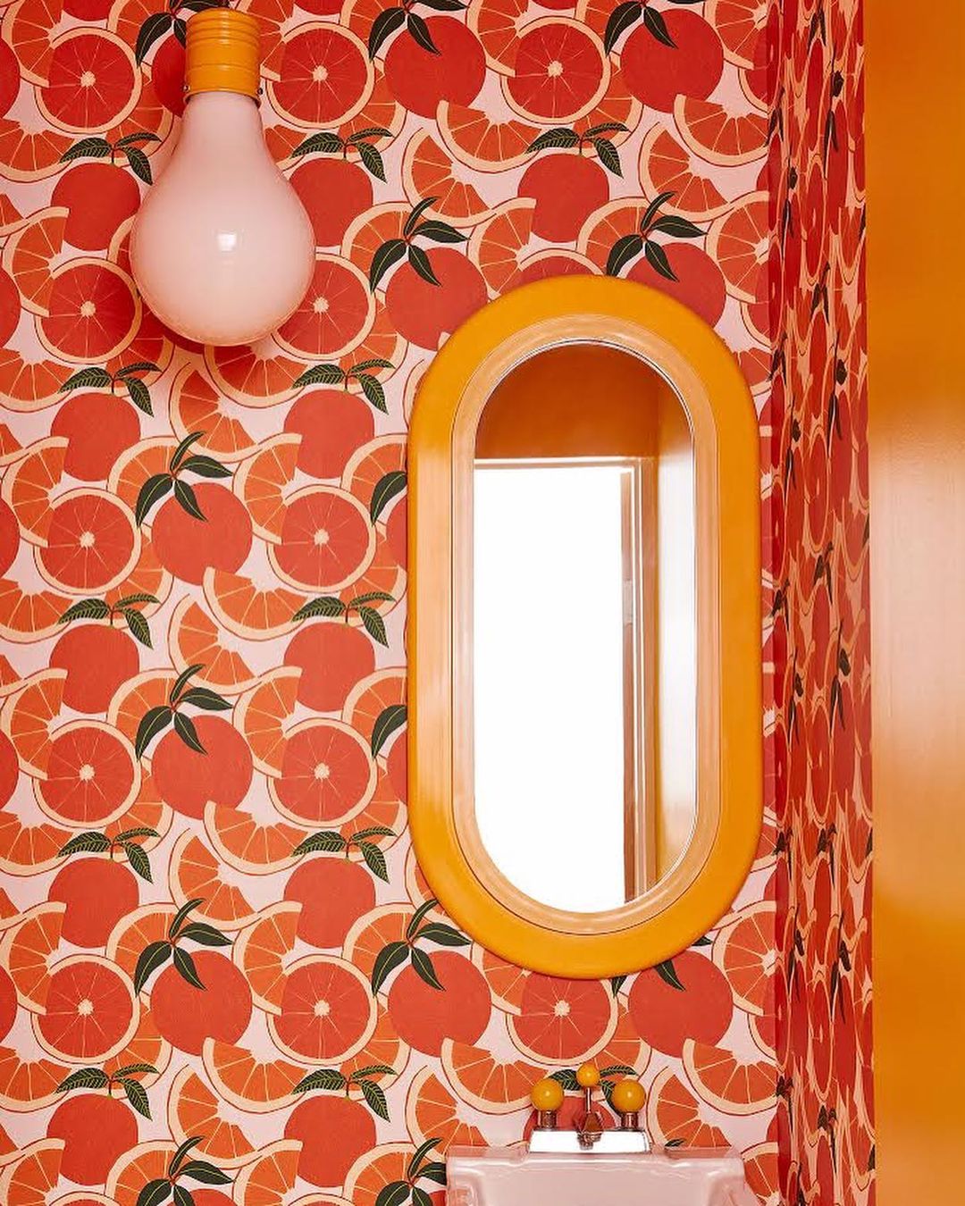 Vibrant bathroom interior with orange-themed wallpaper, illuminated lightbulb, and an oval mirror, creating a retro and inviting space.
