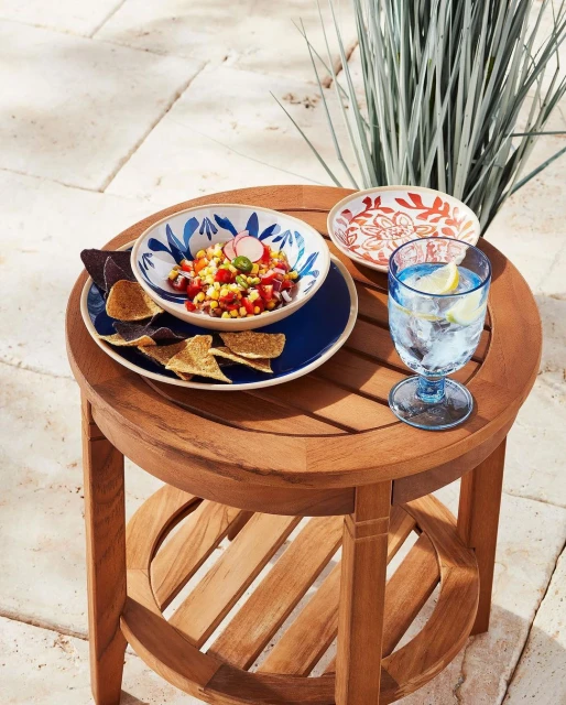 Teak side table with bowl of corn salsa, chips, and a glass of water, styled for outdoor entertaining on a tiled patio.