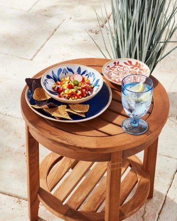 Teak side table with bowl of corn salsa, chips, and a glass of water, styled for outdoor entertaining on a tiled patio.