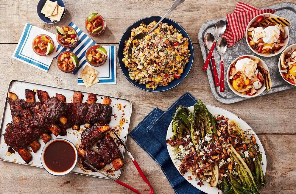 Overhead shot of a wooden table laden with various plates and bowls of colorful grilled food, including ribs, corn salad, and fruit desserts.