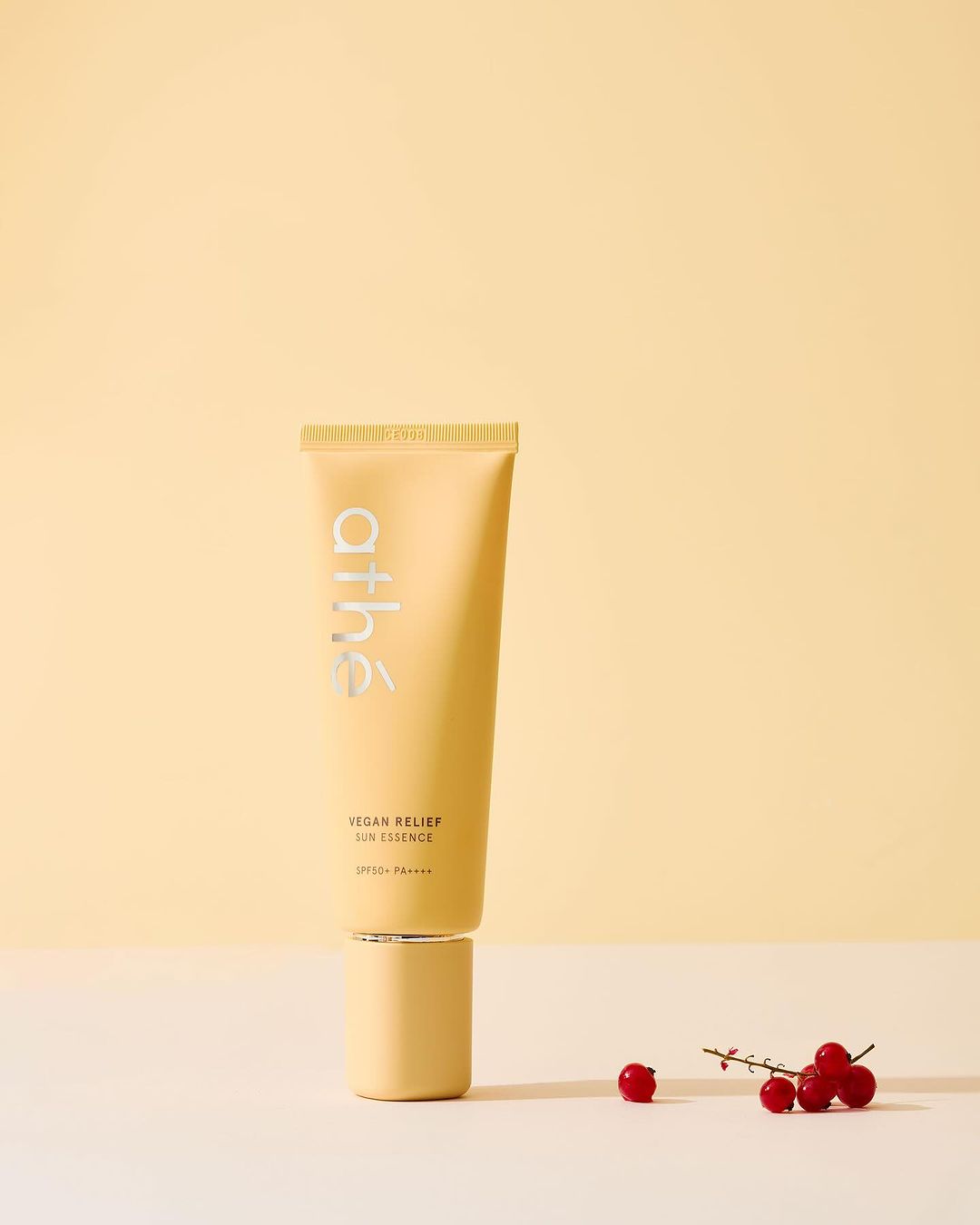 A beige tube of Athé Vegan Relief Lip Essence on a matching pedestal with red currant berries against a yellow backdrop.