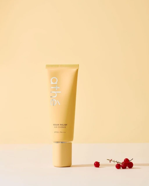 A beige tube of Athé Vegan Relief Lip Essence on a matching pedestal with red currant berries against a yellow backdrop.