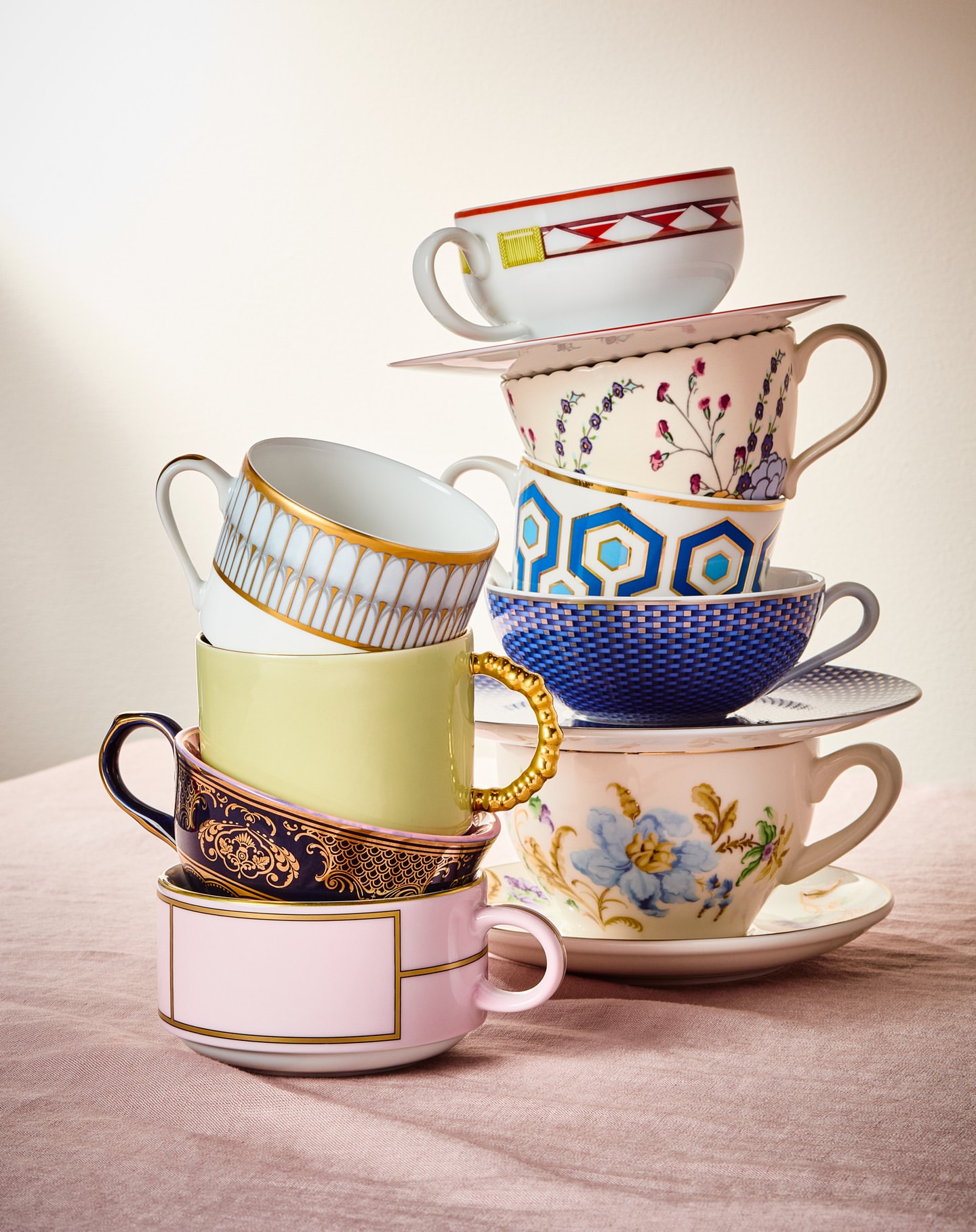Stacked teacups of different colors and patterns, creating a whimsical tower against a neutral backdrop on a light pink textured surface.
