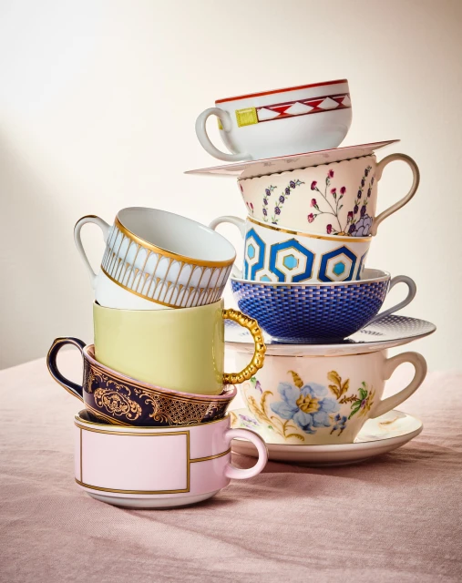 Stacked teacups of different colors and patterns, creating a whimsical tower against a neutral backdrop on a light pink textured surface.