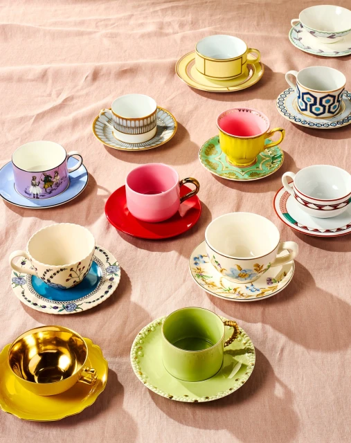 Overhead view of colorful mismatched teacups and saucers arranged on a pale pink tablecloth, showcasing a vibrant collection of ceramics.