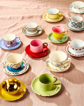Overhead view of colorful mismatched teacups and saucers arranged on a pale pink tablecloth, showcasing a vibrant collection of ceramics.