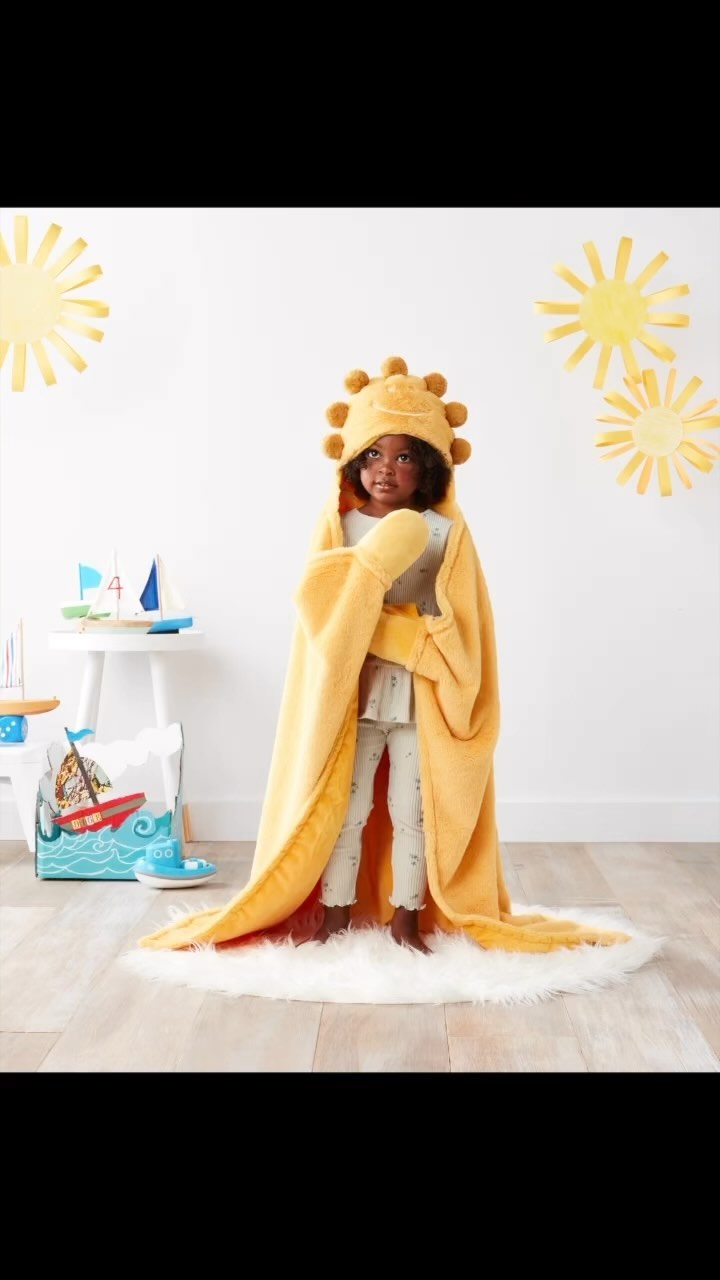 A child stands on a white rug, wrapped in a yellow hooded sun-themed towel, in a light and airy indoor setting with nautical decor.