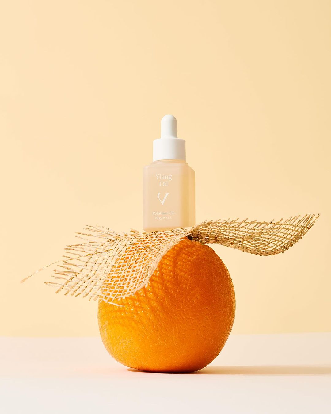 Close-up shot of a skincare oil bottle resting on an orange, accented with tan netting against a soft yellow background.