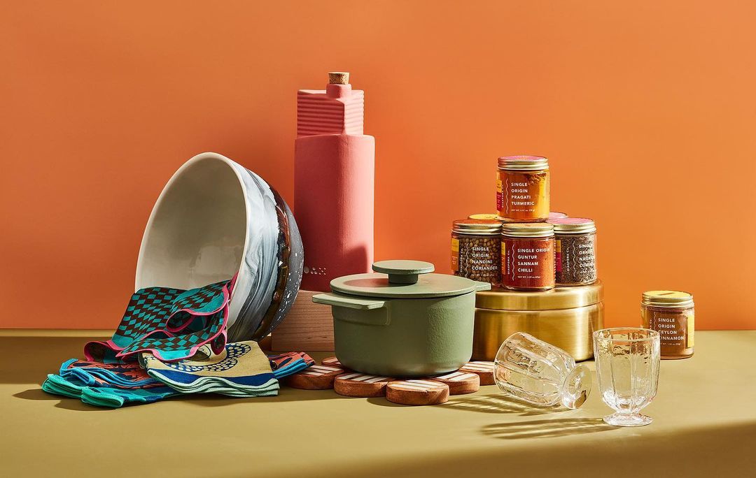 Still life shot of a green pot, patterned textiles, glassware, spice jars, and abstract decorative items against an orange backdrop.