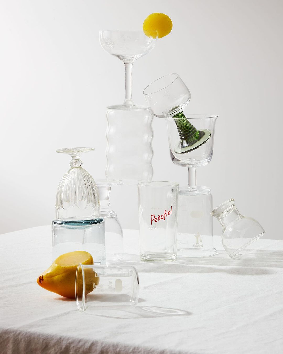 Still life studio photography of assorted glassware stacked on a white tablecloth with two lemons, creating a minimalist, bright composition.