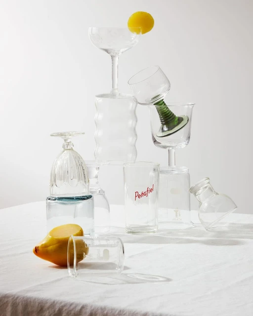 Still life studio photography of assorted glassware stacked on a white tablecloth with two lemons, creating a minimalist, bright composition.