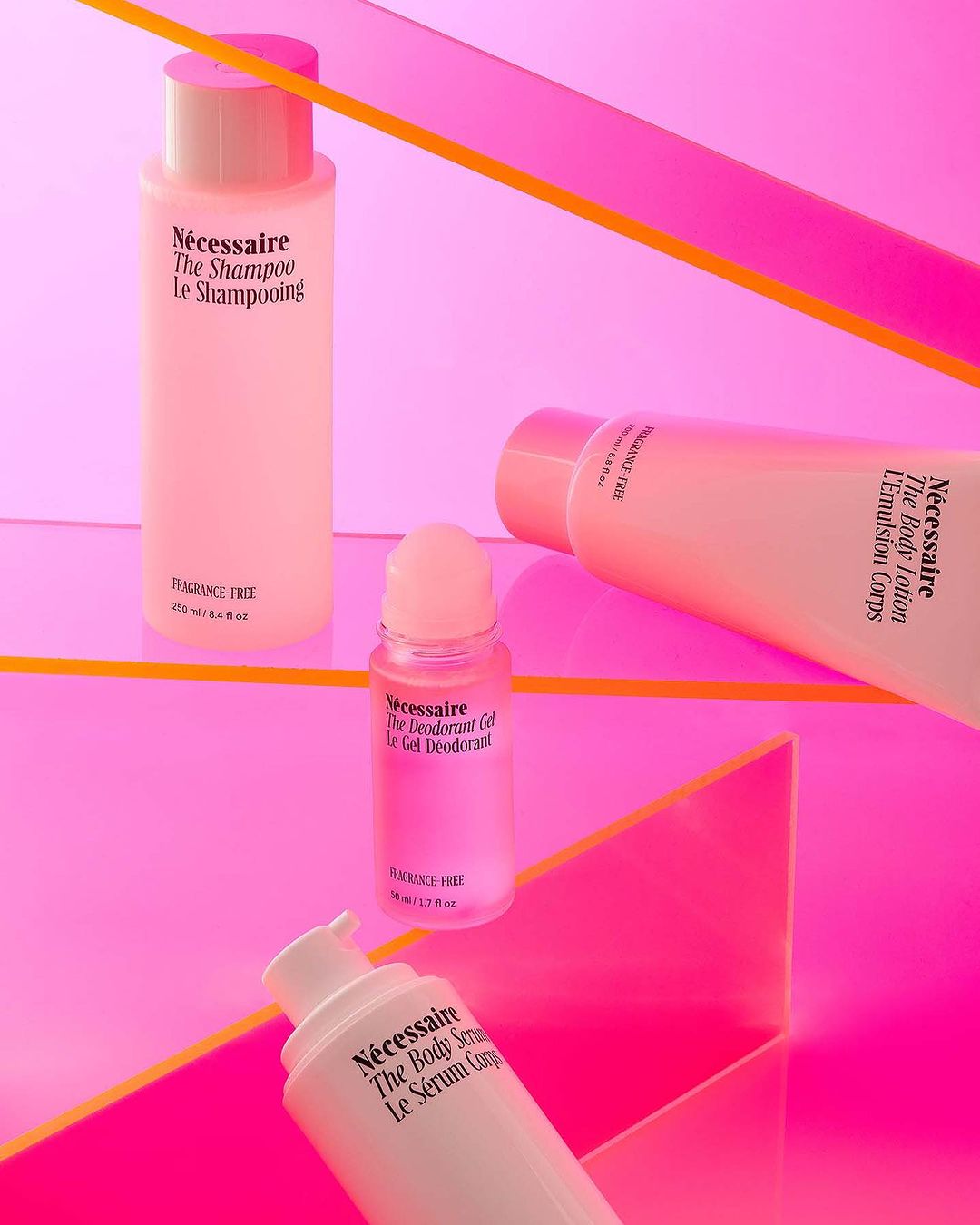 Product photography of Nécessaire brand body wash, shampoo, lotion, and deodorant in pink and white bottles against a pink backdrop.