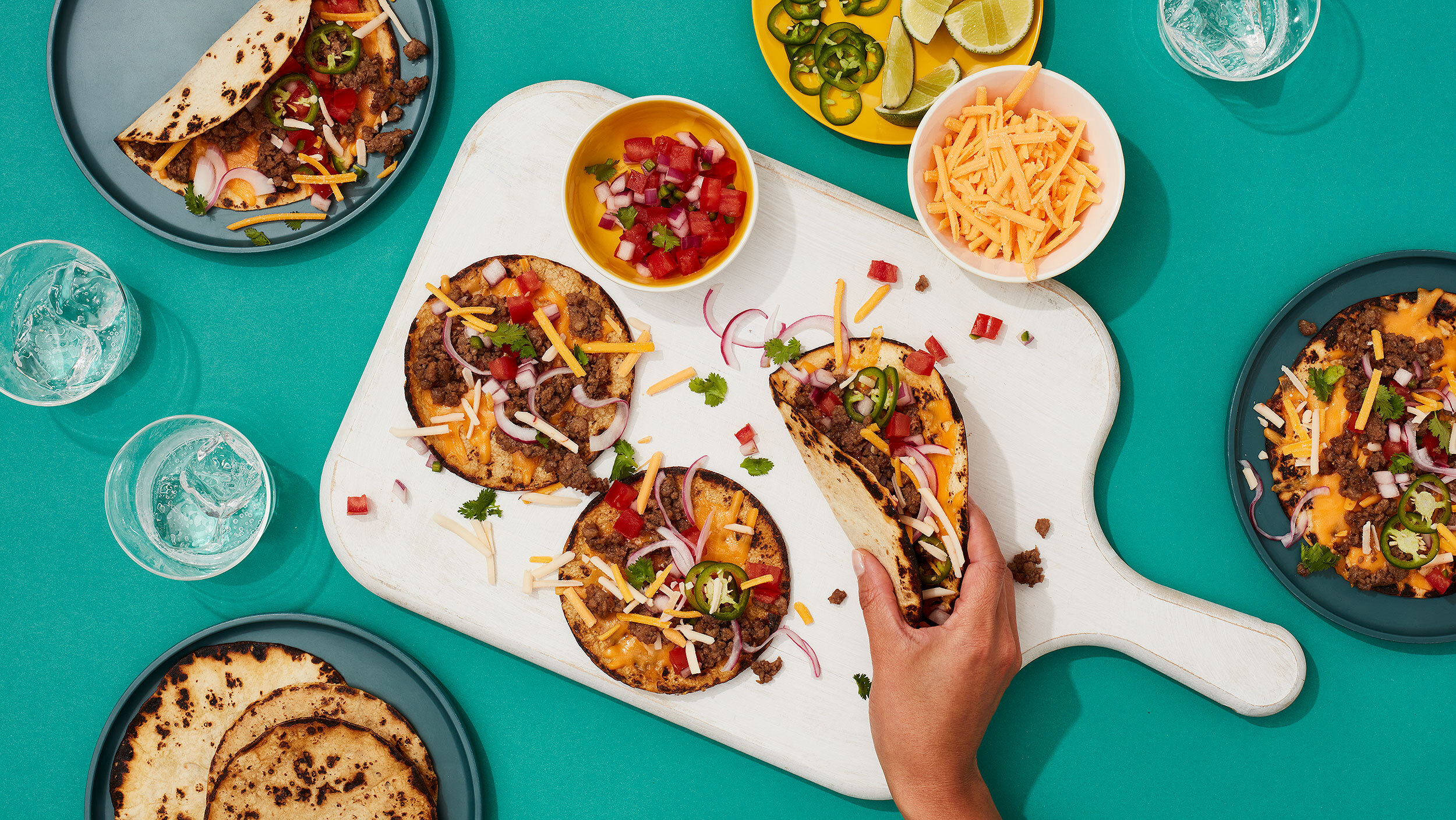 A hand holds a taco over a white wooden board topped with tacos, bowls of shredded cheese, pico de gallo, lime, and jalapeños on a teal surface.