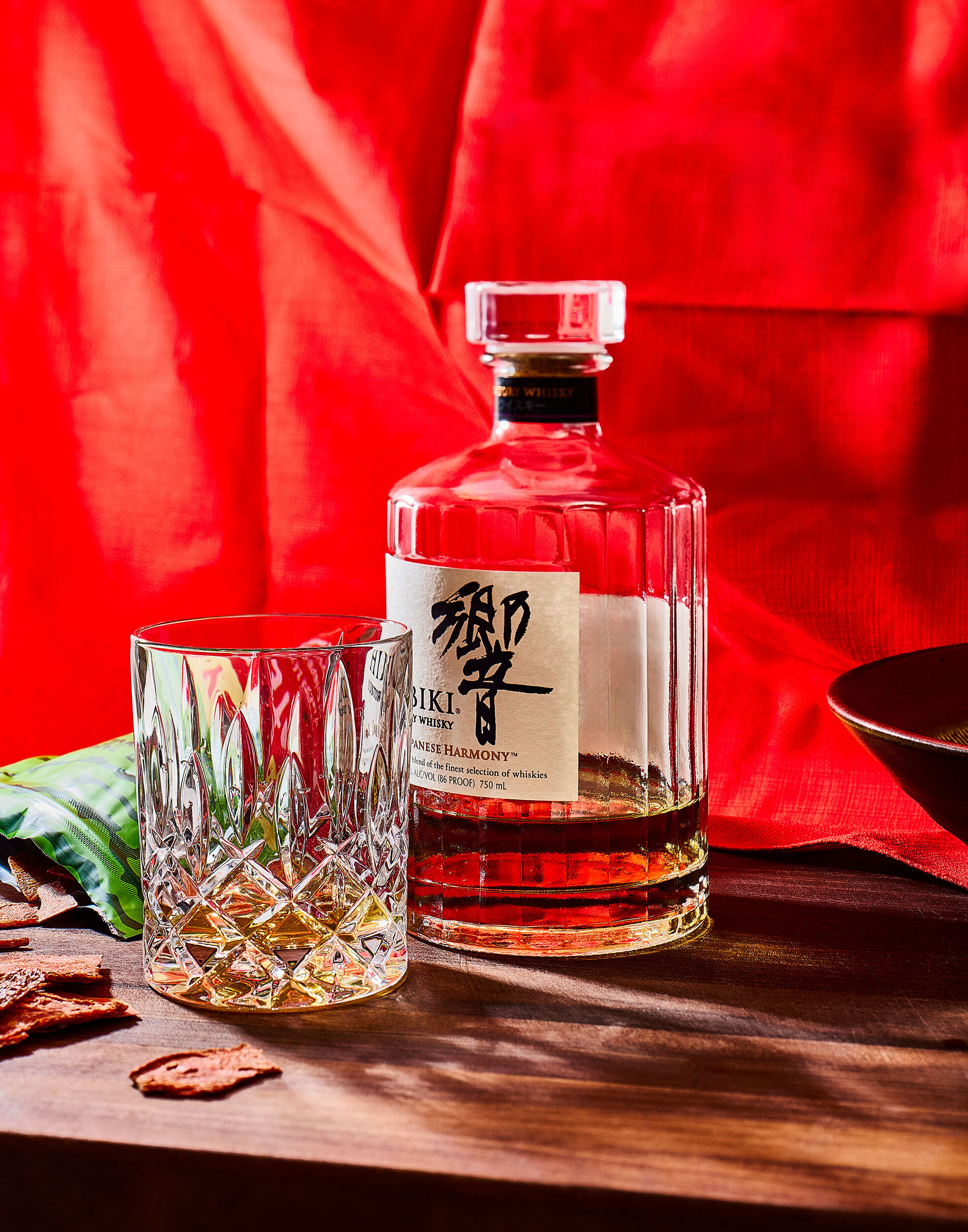 A crystal whiskey glass and a decanter of Hibiki Japanese Harmony whisky on a wooden surface against a vibrant, draped red fabric background.