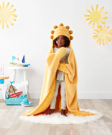 Young child in a plush yellow hooded sun blanket stands on a fuzzy rug in a brightly lit studio with nautical-themed paper decor.