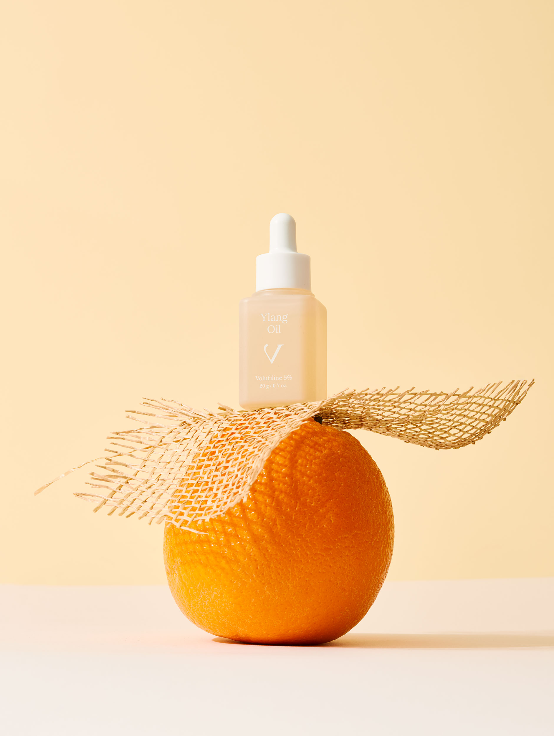 A small glass dropper bottle of Ylang Oil resting atop a whole orange, with a strip of burlap textured fabric wrapped around the base.