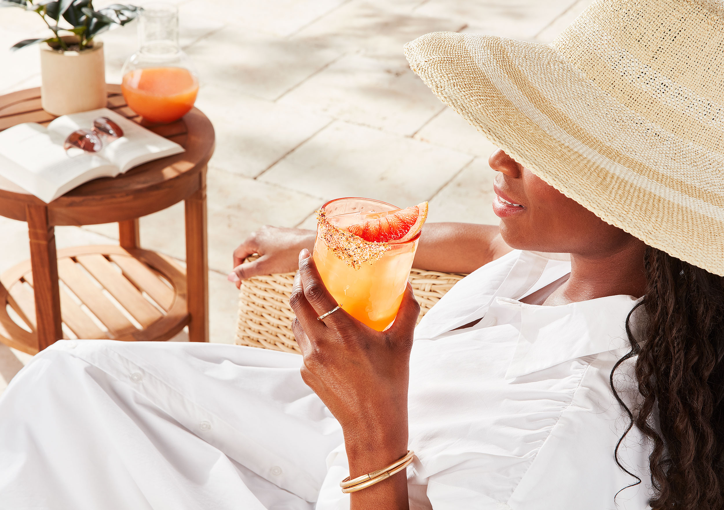 A woman wearing a straw hat and white dress holding a citrus cocktail garnished with a grapefruit slice outdoors.