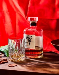 A bottle of Hibiki Japanese Harmony whisky and a cut crystal tumbler set on a wooden surface against draped red fabric.