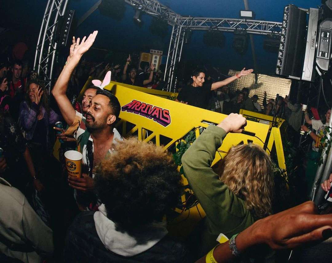 Crowd enjoys a DJ performance at a music event; performer controls the music behind a yellow soundboard with crowd in foreground.