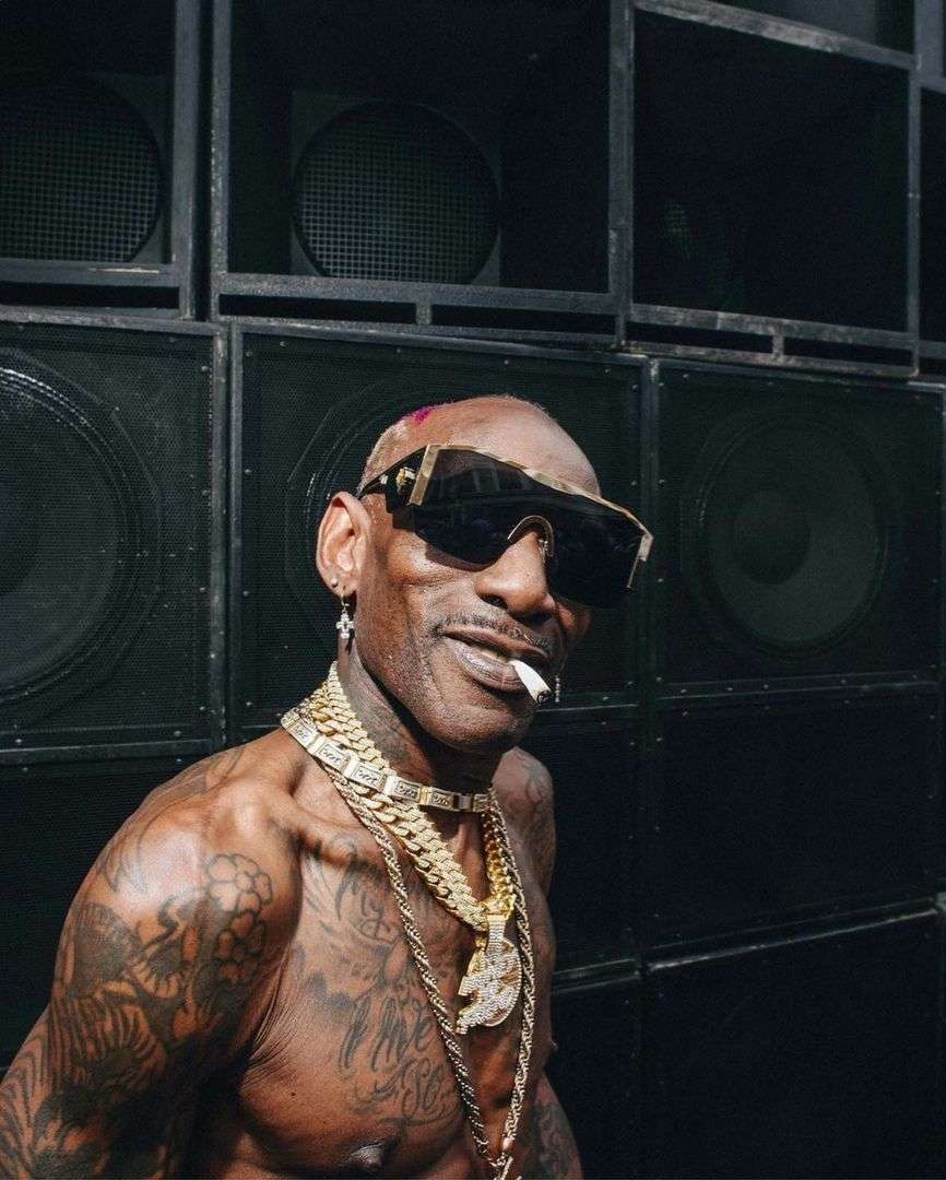 Adult man with tattoos and gold chains posing in front of a wall of speakers, wearing sunglasses and smoking a cigarette.