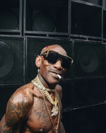 Adult man with tattoos and gold chains posing in front of a wall of speakers, wearing sunglasses and smoking a cigarette.