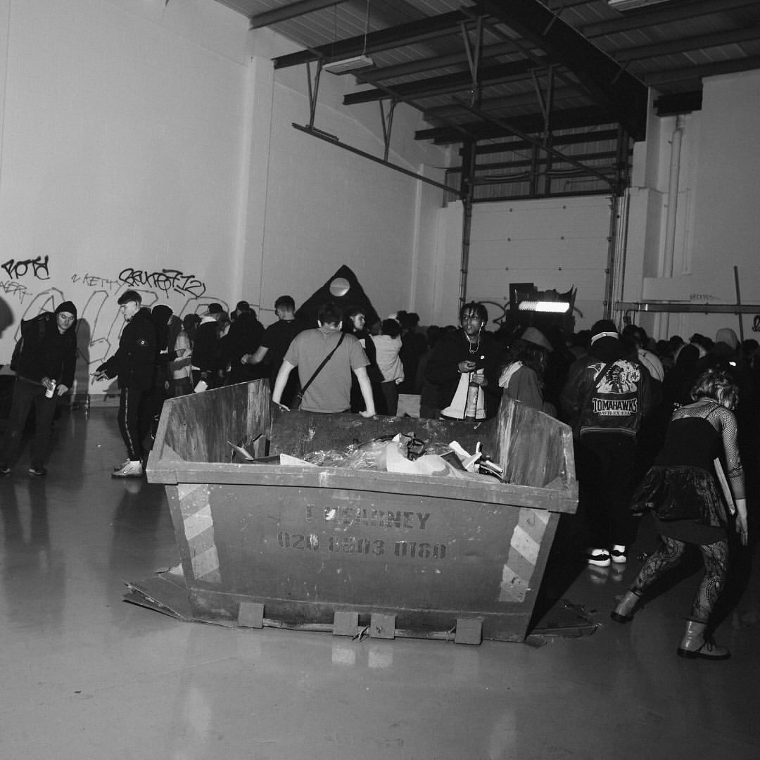 Monochrome shot of a large group of people in an indoor space, with a dumpster overflowing with debris in the foreground.