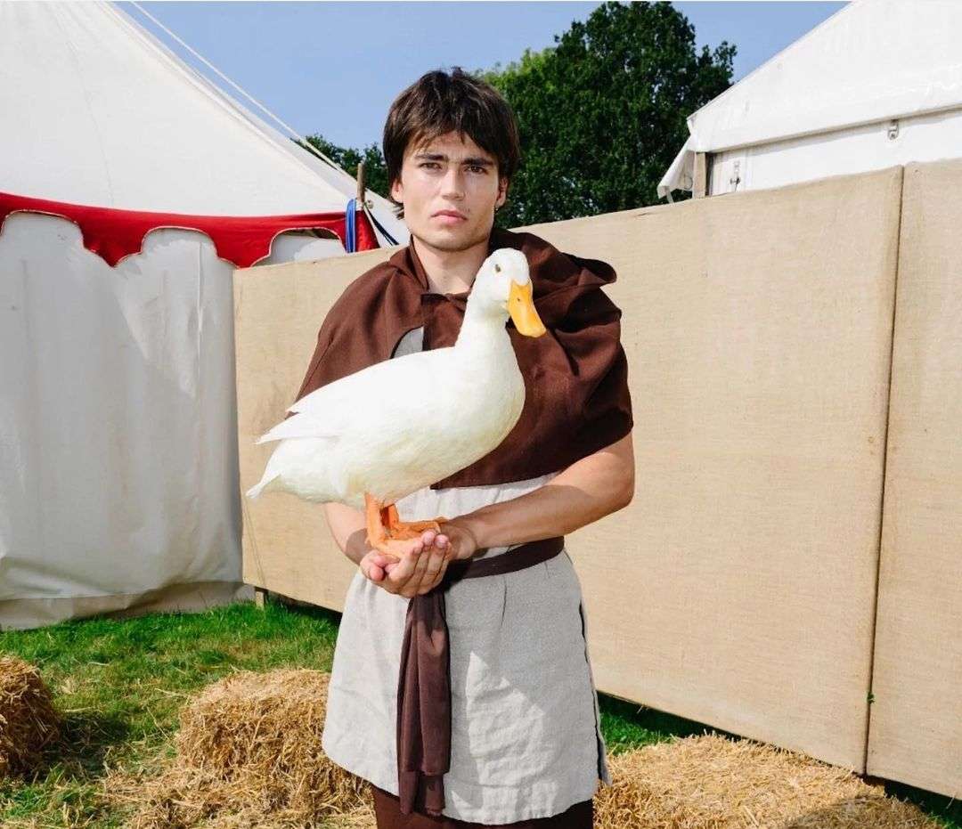Young man in medieval-style clothing holds a white duck in an outdoor setting with tents and hay bales, creating a unique portrait.