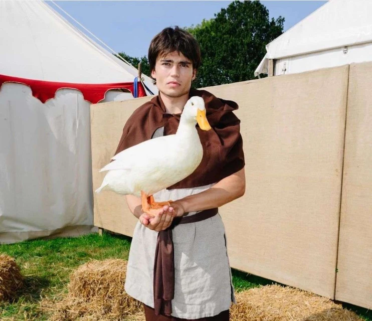 Young man in medieval-style clothing holds a white duck in an outdoor setting with tents and hay bales, creating a unique portrait.