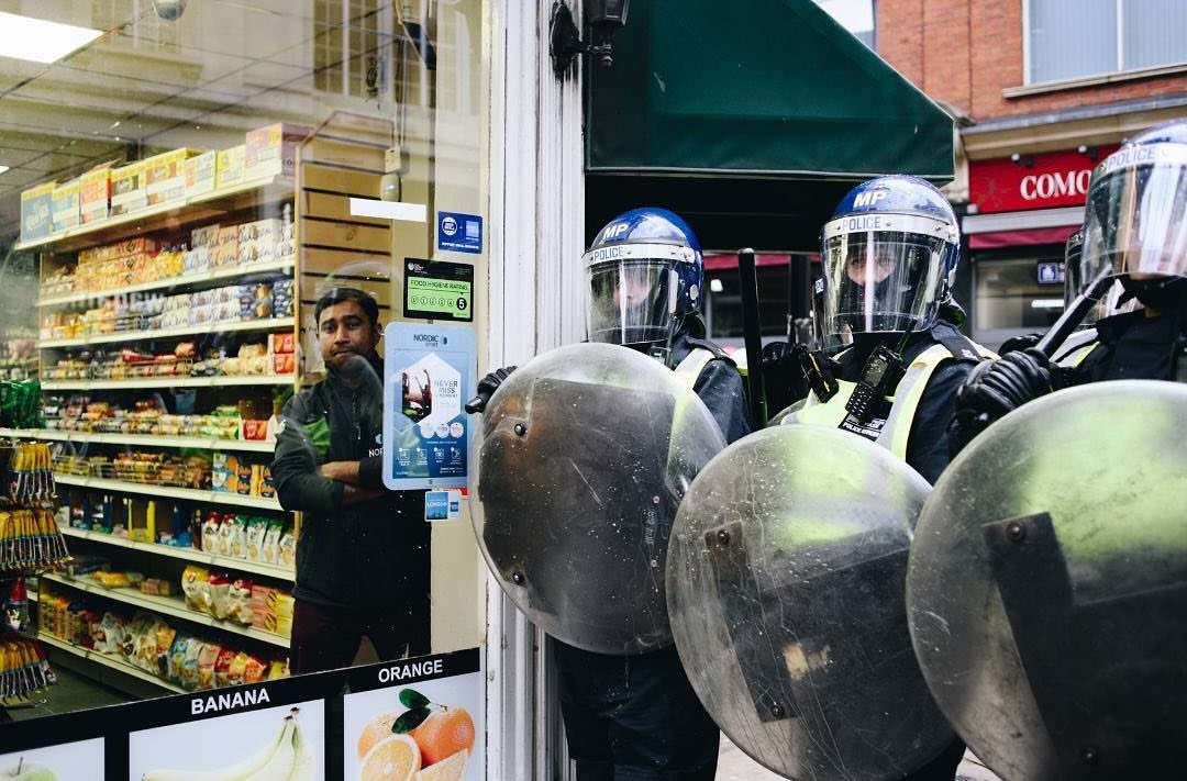 Police officers in riot gear stand guard in front of a storefront, a man inside the store observes the scene. Urban setting, daytime.