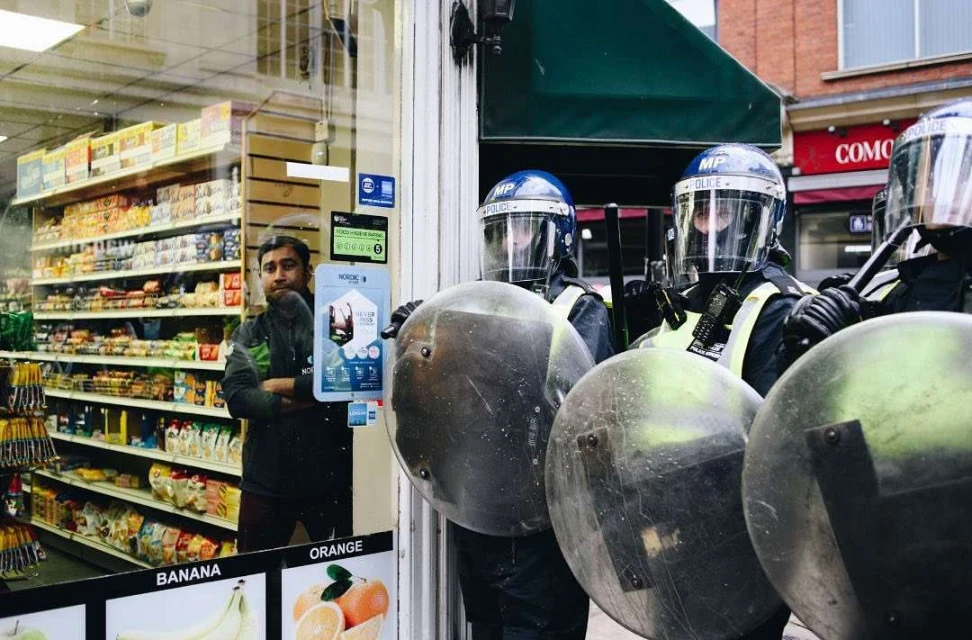 Police officers in riot gear stand guard in front of a storefront, a man inside the store observes the scene. Urban setting, daytime.