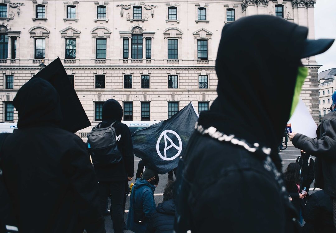 People gather in an urban environment, some wearing hoods and a mask, with a banner featuring a white symbol in a circle.