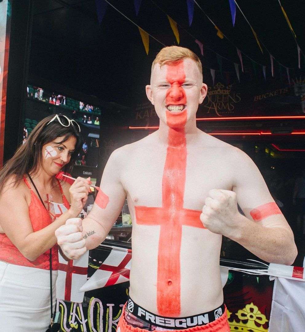 Enthusiastic fair-skinned man with a painted England flag on his torso poses with clenched fists and a woman painting the cheek of another woman.
