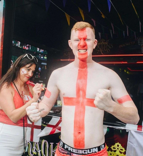 Enthusiastic fair-skinned man with a painted England flag on his torso poses with clenched fists and a woman painting the cheek of another woman.