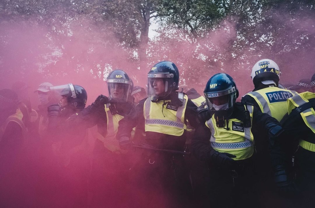 Group of police officers wearing riot gear stand in a line amidst purple smoke in an outdoor setting, focusing on law enforcement.