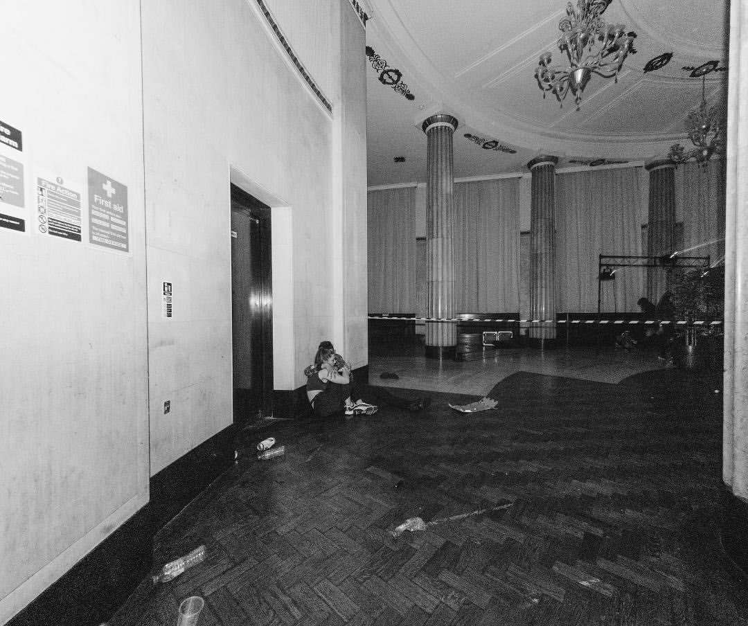 Black-and-white image of a woman sitting on the floor in a large, empty hall with columns and a chandelier, near signage on the wall.