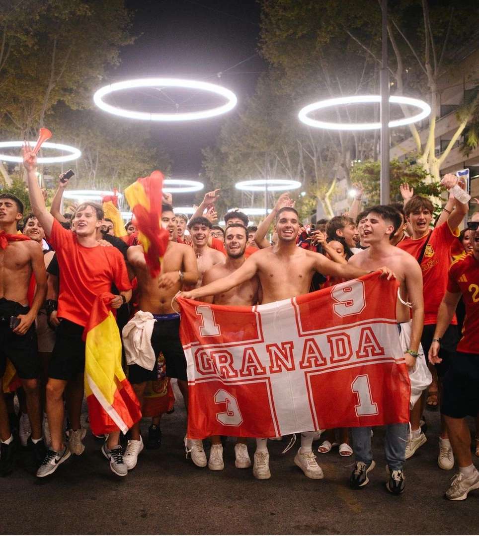 Enthusiastic group of young adult men cheer in a city street at night holding a sports team flag; illuminated rings overhead.