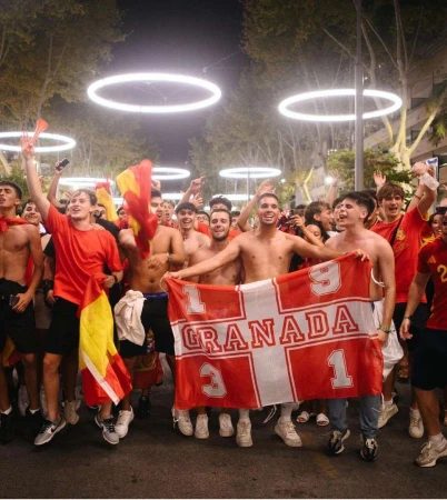 Enthusiastic group of young adult men cheer in a city street at night holding a sports team flag; illuminated rings overhead.