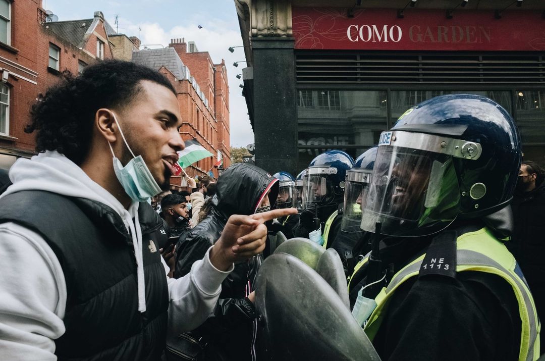 A protester gestures towards police officers wearing riot gear on a city street with buildings and signage visible in the background.