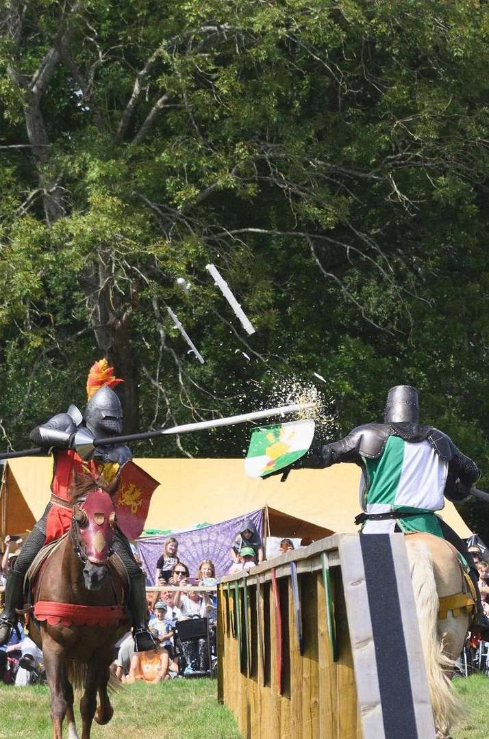 Two knights on horseback jousting in an outdoor arena, wearing armor and helmets, with a crowd watching in the background on a sunny day.