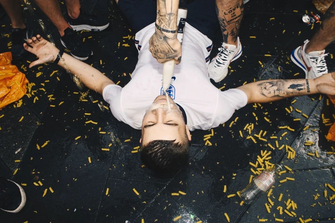 Man in soccer jersey lying on a black surface, receiving a liquid from a bottle; surrounding people and scattered pasta add to the scene.