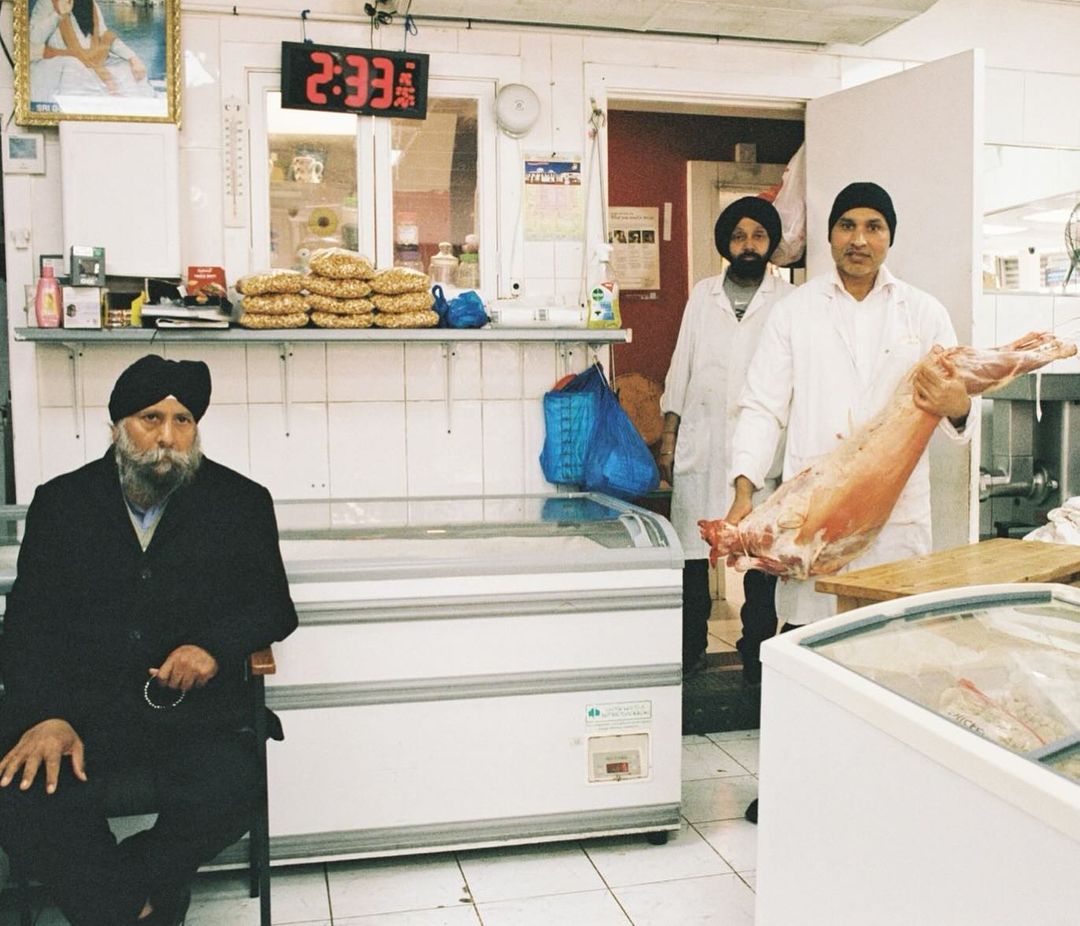 Two Sikh men in white coats hold a large cut of meat in a market. A third man sits in foreground with digital clock in background.