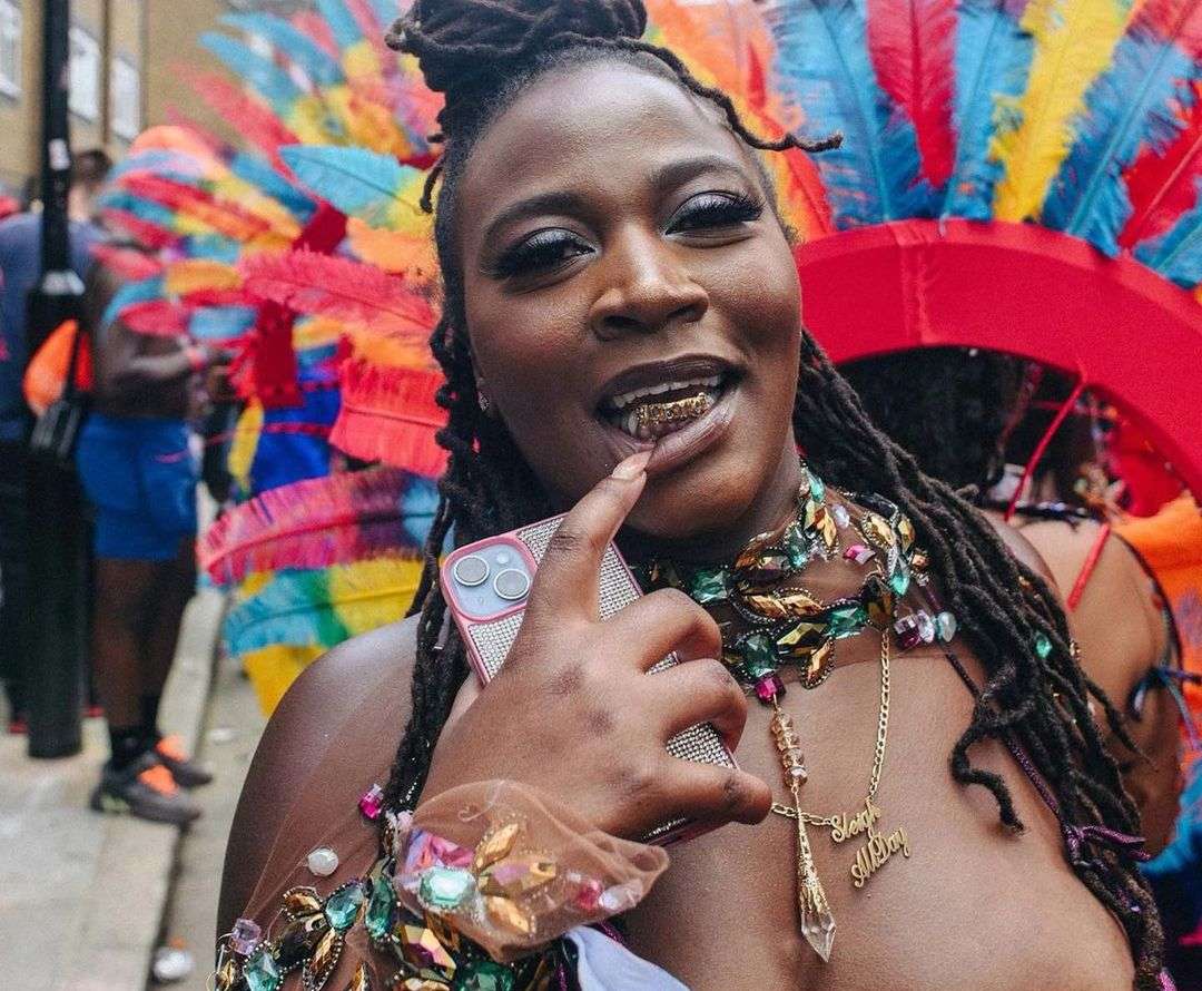 Woman with dreadlocks and gold teeth, wearing a beaded top and necklace, poses with a phone at a colorful outdoor event.