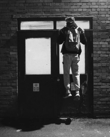 Monochrome photograph of a person hanging from the top of a dark double door against a brick wall at night by Yushy Pachnanda.