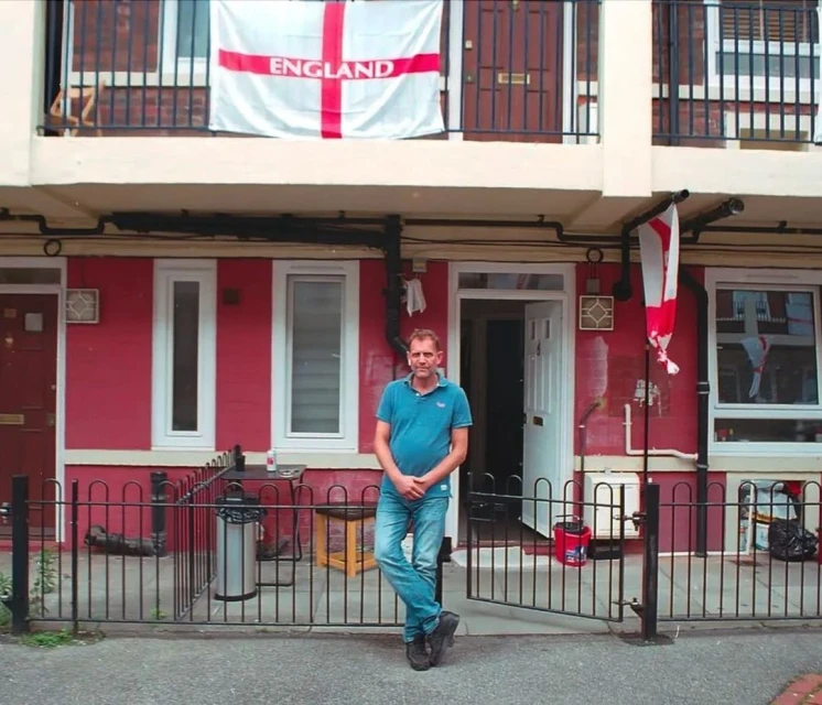 Adult male standing in front of red brick apartments with England flags, creating an urban, patriotic scene with casual day lighting.