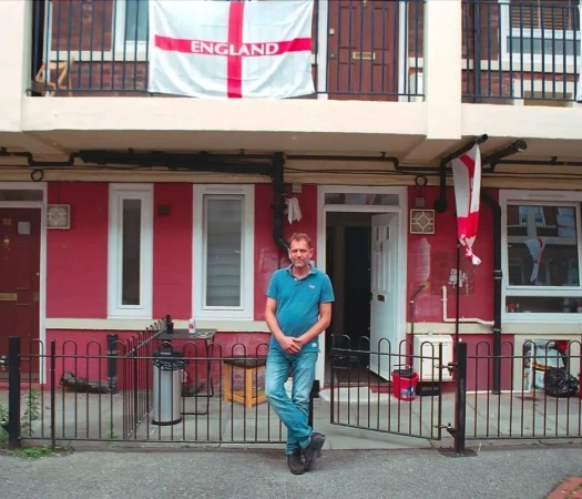 Adult male standing in front of red brick apartments with England flags, creating an urban, patriotic scene with casual day lighting.