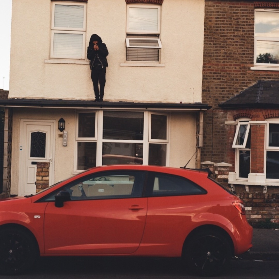 A person in dark clothing stands on the ledge of a two-story house next to a red compact car on a street in daylight.