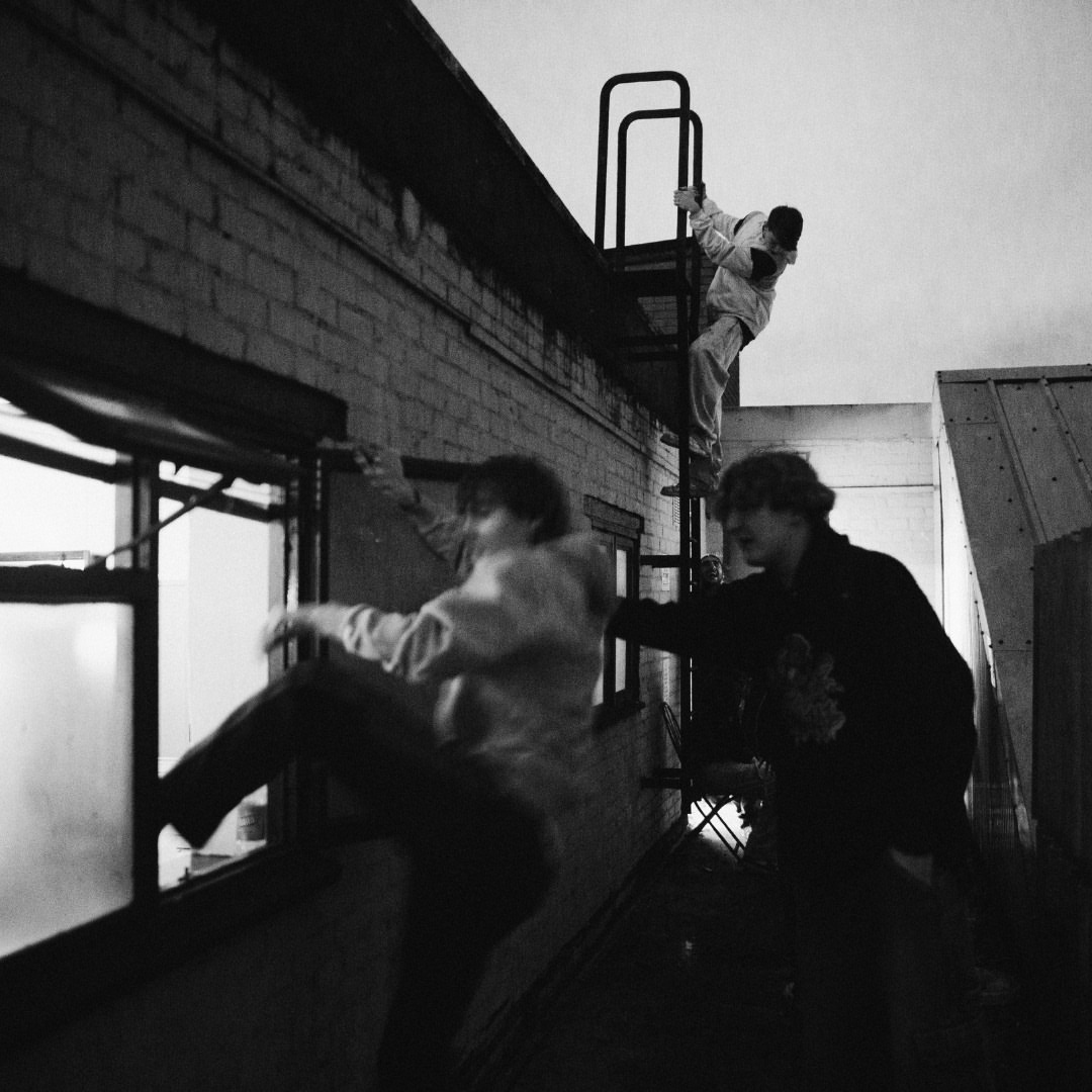 Three people are climbing a building; one person is climbing a ladder, and two people are at a window, with a cloudy sky in the background.