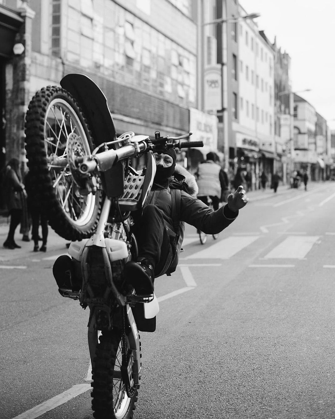 Black and white photo of a person on a dirt bike doing a wheelie on a city street lined with buildings and pedestrians in the background.